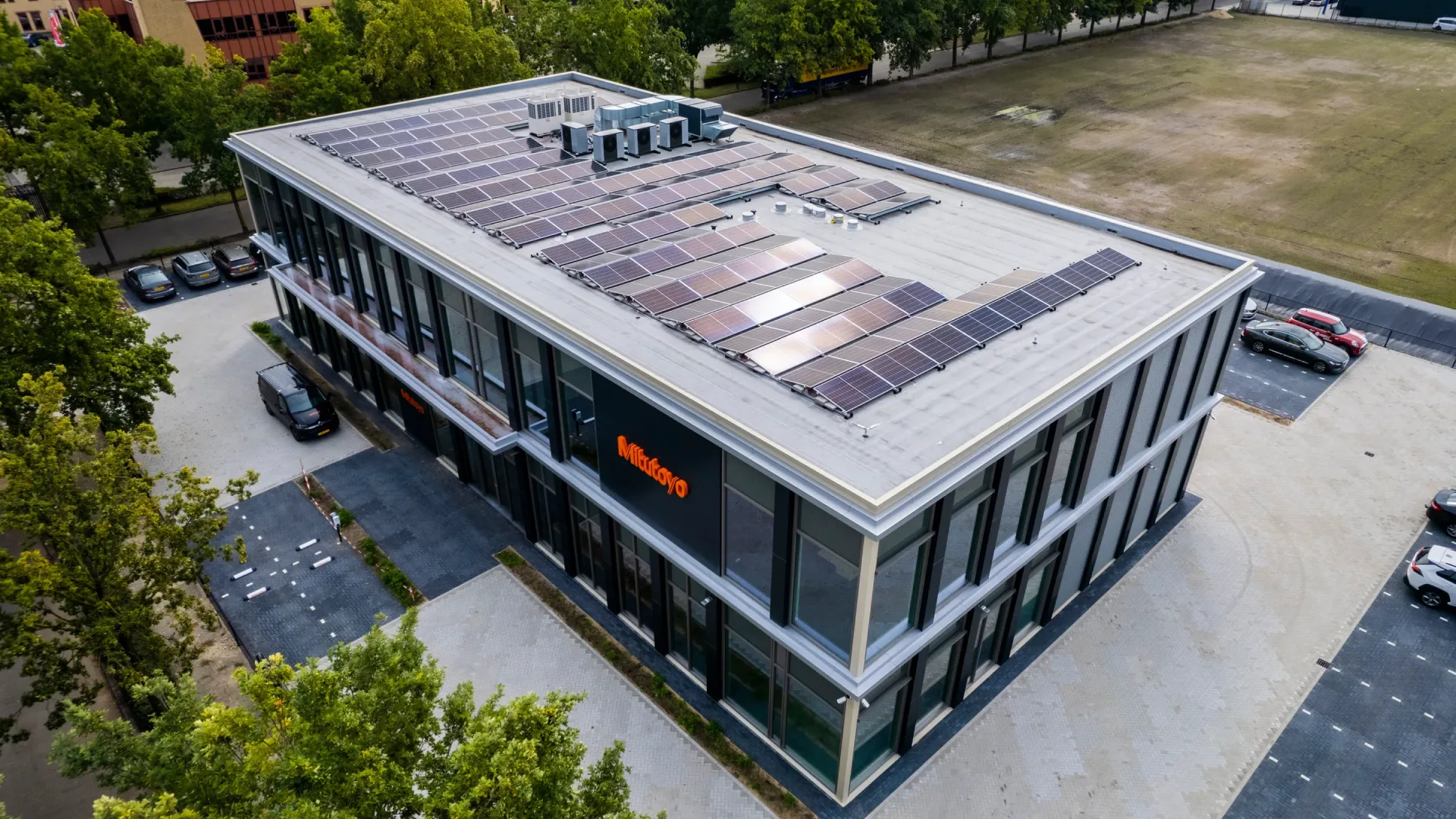 Luftaufnahme eines Mitutoyo-Bürogebäudes mit einer großen Anzahl von Solarpanels auf dem Dach, einem Parkplatz und umgebenden Bäumen.