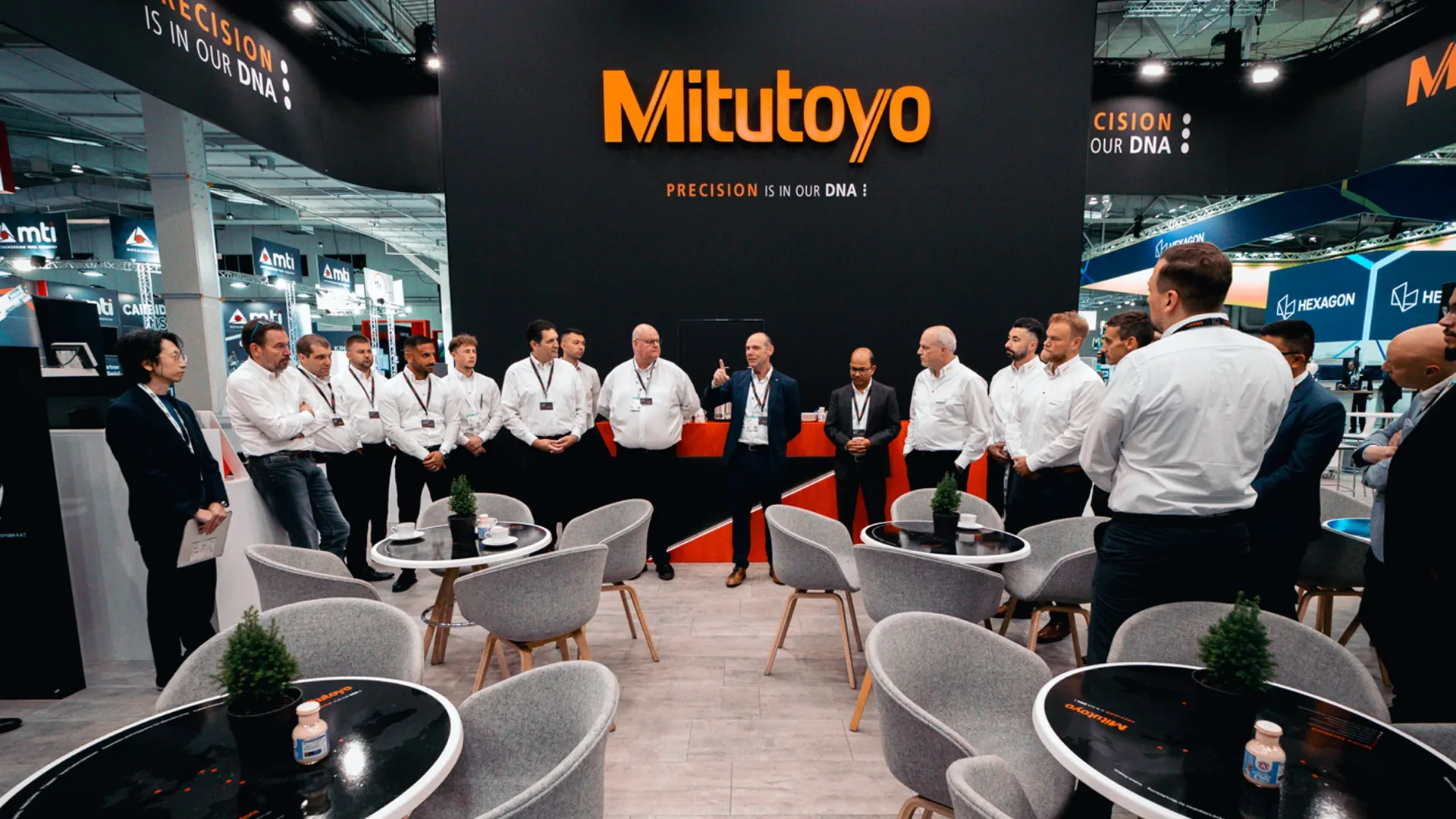 Gruppe von Mitutoyo-Mitarbeitern in Unternehmenskleidung, die sich für ein Briefing an einem Messestand versammelt haben; Mitutoyo-Branding und der Slogan „PRÄZISION IST IN UNSERER DNA“ sind hinter ihnen sichtbar.